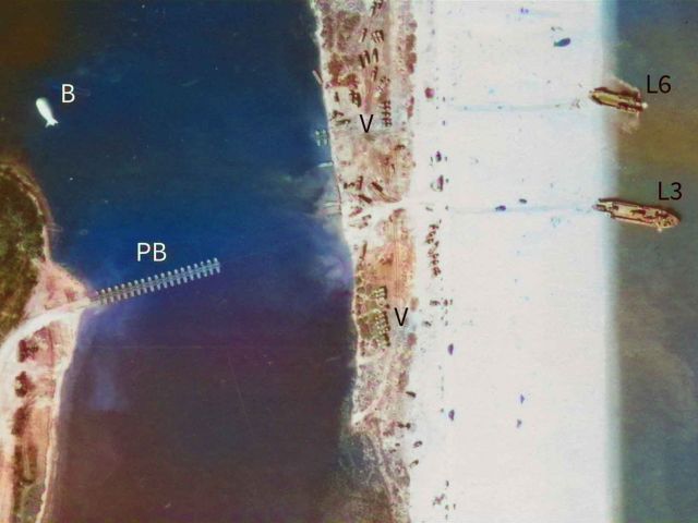 Detail from usaaf-022-us-30gr-loc41-v-0035 aerial photograph, showing a pontoon bridge being constructed on a stretch of water between a landward road and a narrow strip of beach. Two landing craft are stationary at the edge of the beach. Vehicles disembark from one of them. Smaller landing craft manoeuvre on the sea close to them. Various vehicles are on the beach and on land between the beach and the pontoon bridge. The letters and numbes 'PB', 'B', 'V', 'L3' and 'L6' applied to the image mark locations.
