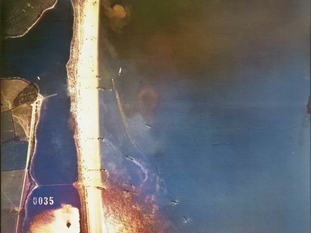 Colourised USAAF aerial view image of Exercise Tiger on 27 April 1944 showing the northern half of the lake behind the beach called Slapton Ley. Various Landing Craft Tanks are arriving at the beach, unloading vehicles or sailing off back into Start Bay. Lines of vehicles can be seen driving up the beach slope to the lakeside. Original Image Source: <a href="https://historicengland.org.uk/images-books/archive/collections/photographs/usaaf-collection-exercise-tiger/" alt="Click this link to visit the Historic England website Witnessing Exercise Tiger: USAAF Photos from April 1944 section with more information about the photographs, in anew browser tab or window." target="_blank">Historic England</a>. Archive, USAAF Photography.